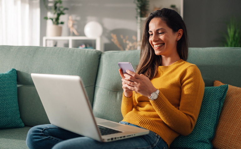 Woman on here smartphone smiling and have a laptop on the knees.