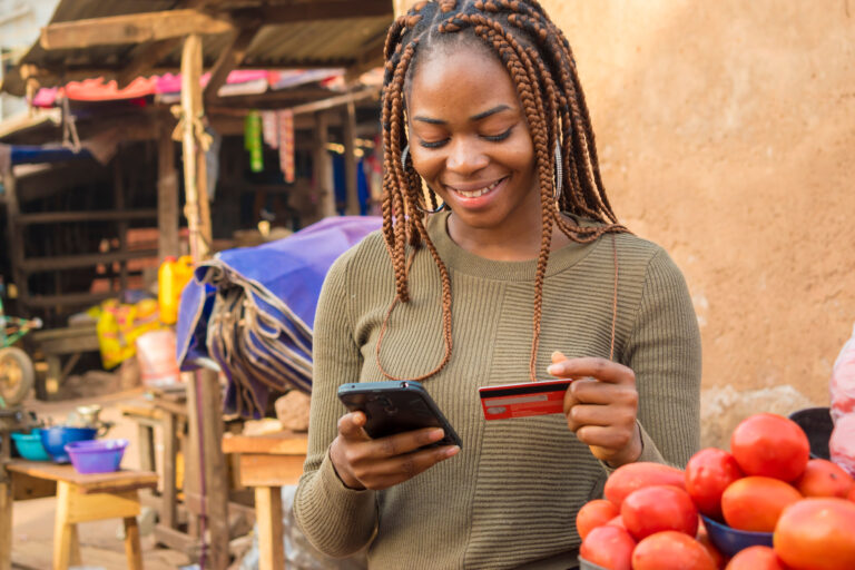 Smiling woman holding a smartphone and a credit card at an outdoor market.